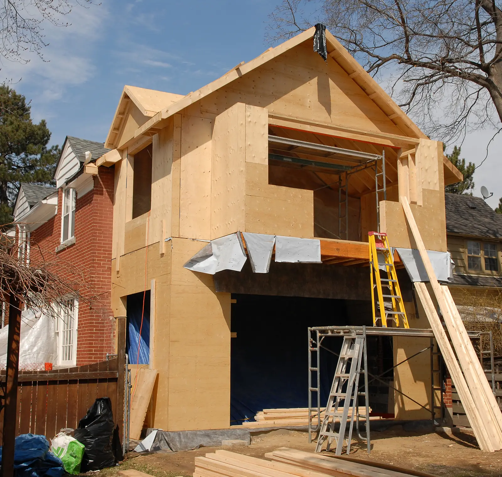 Two-story house under construction with wooden framing. Scaffolding and ladders are positioned at the front. Brick house is visible to the left.