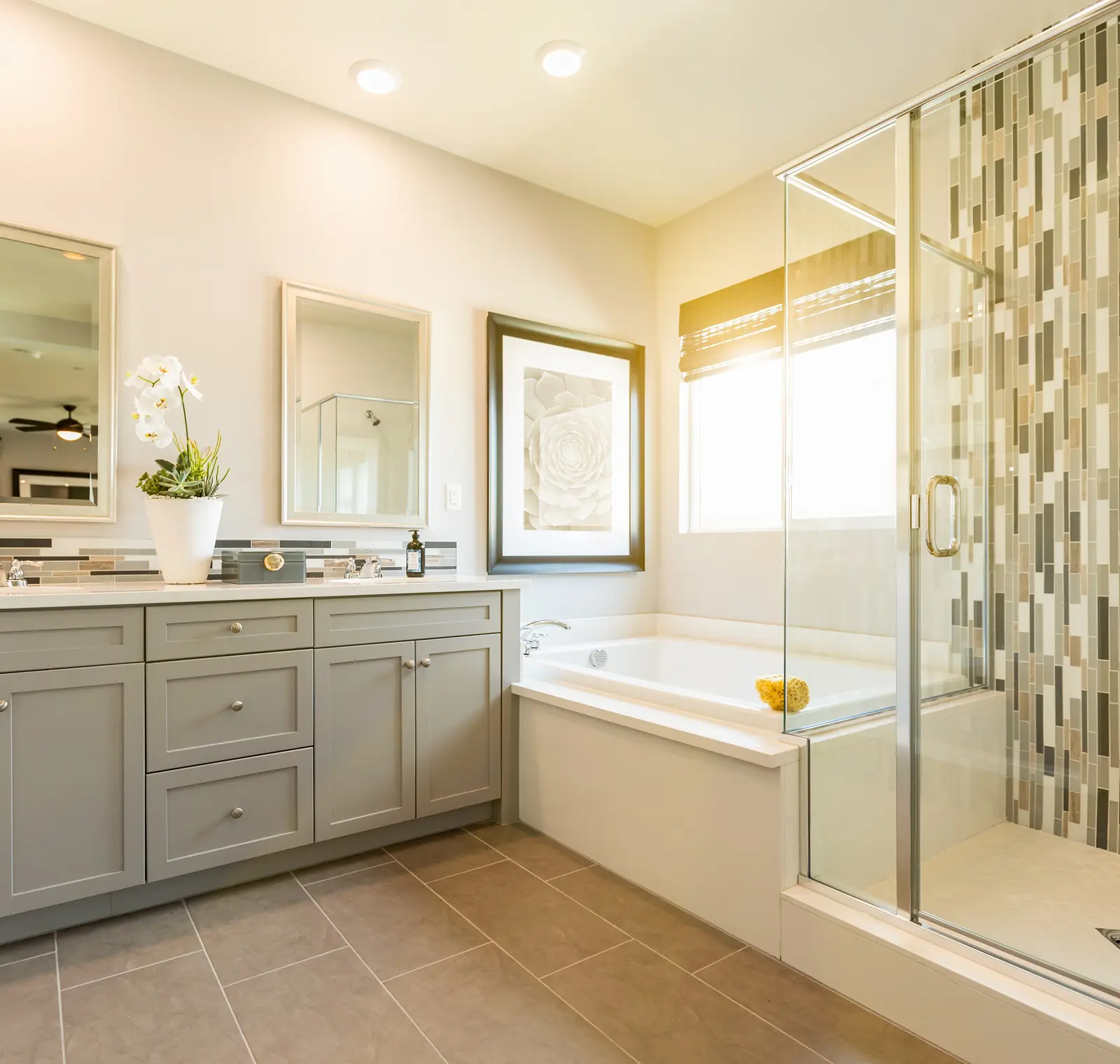 Modern bathroom with grey cabinets, white countertops, potted orchid, framed mirror and artwork, glass shower enclosure, and a bathtub by the window with patterned tile backsplash.