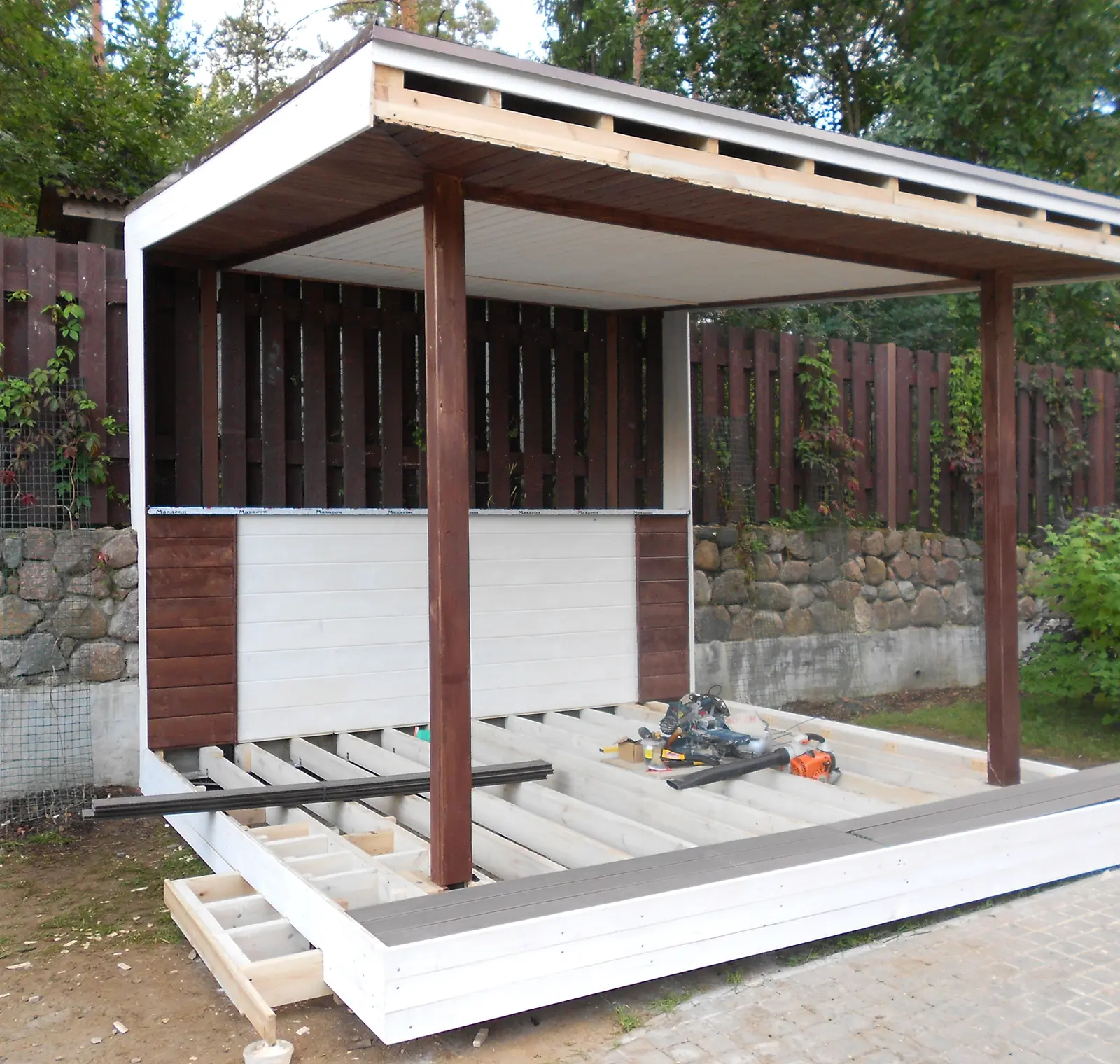 Wooden gazebo under construction, featuring a roof, wooden frame, and partially completed floor. Garden tools are placed on the floor panels. Background shows a wooden fence and greenery.