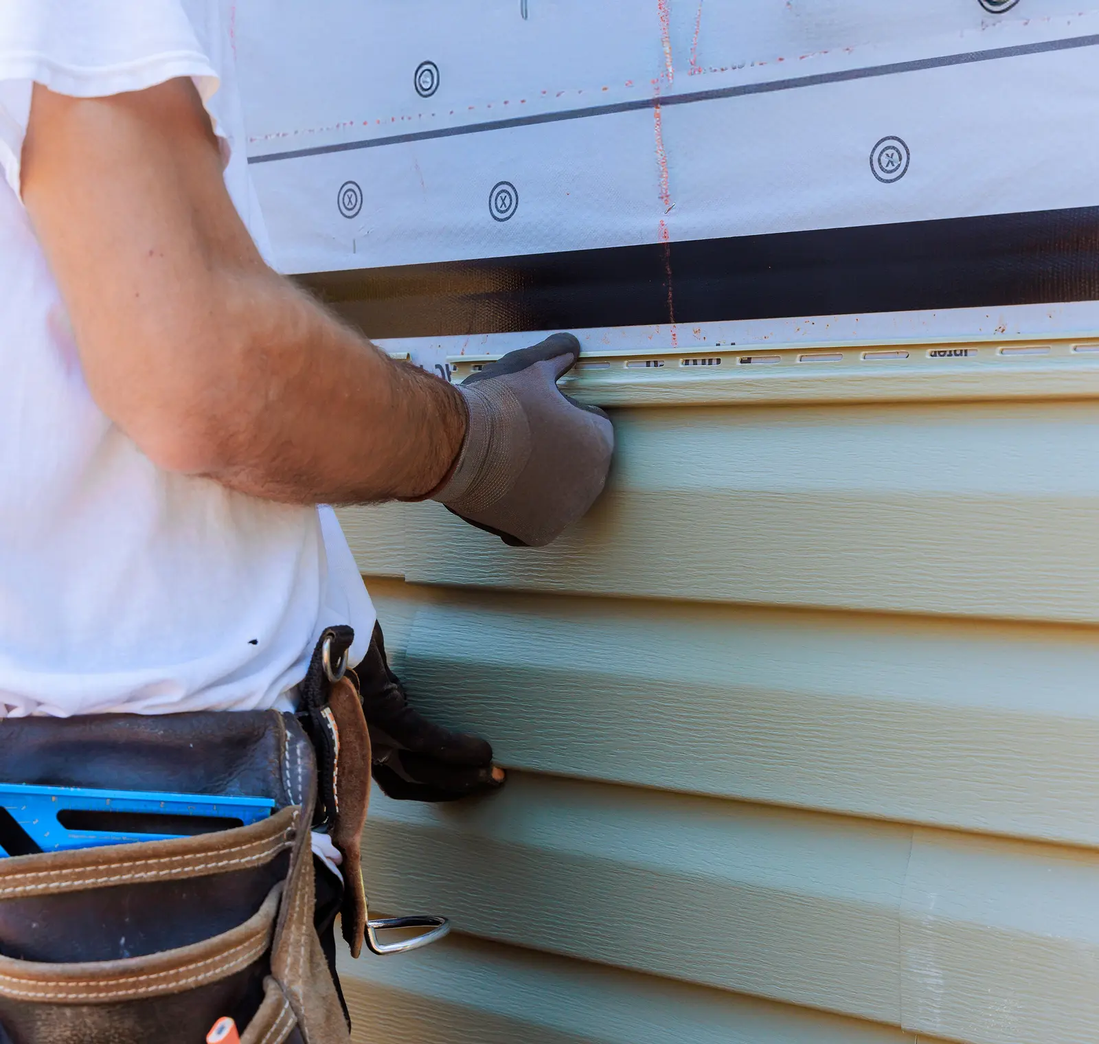 Person wearing a tool belt is installing light green vinyl siding on a house, using a level for alignment.