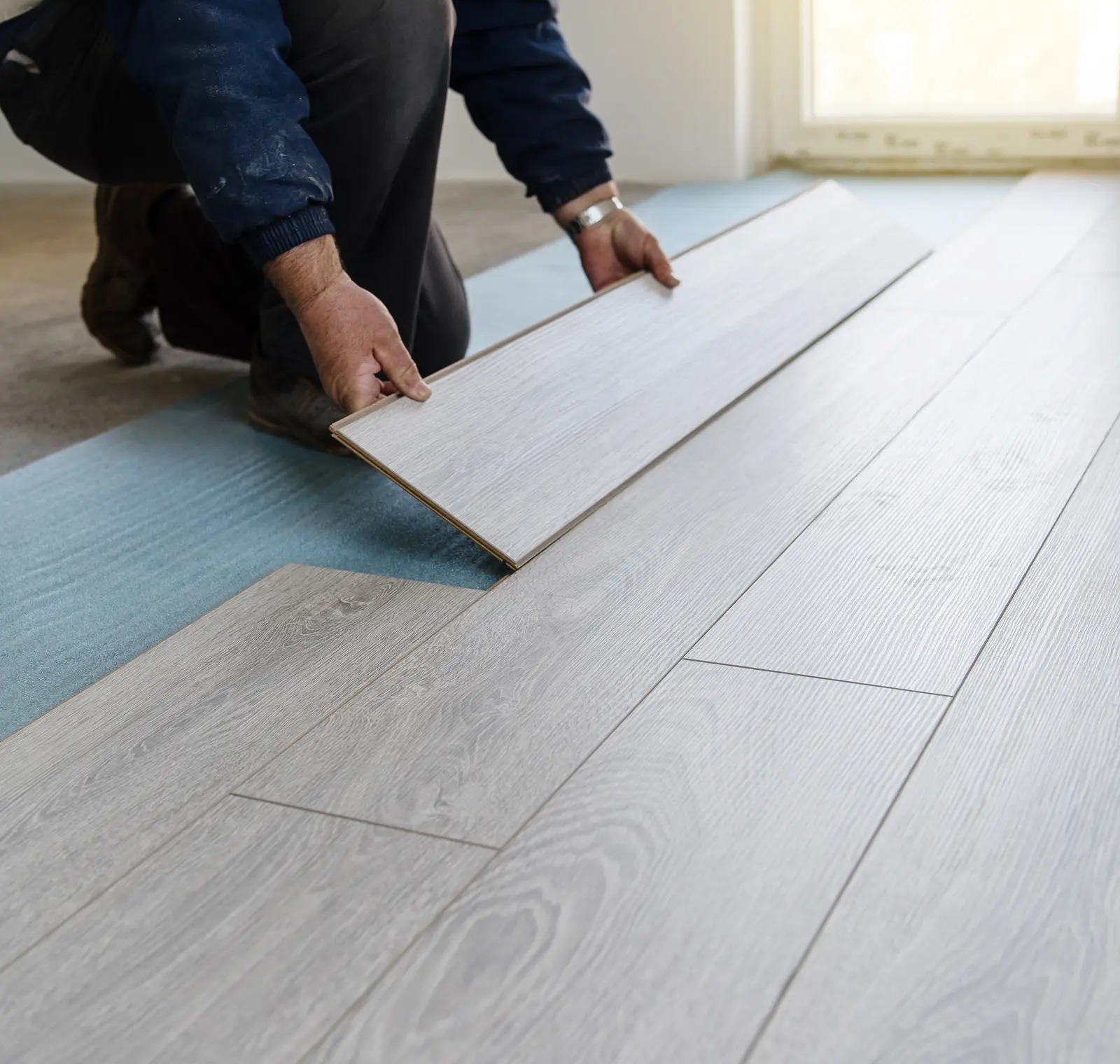 Person installing light gray wood laminate flooring over a blue underlay.