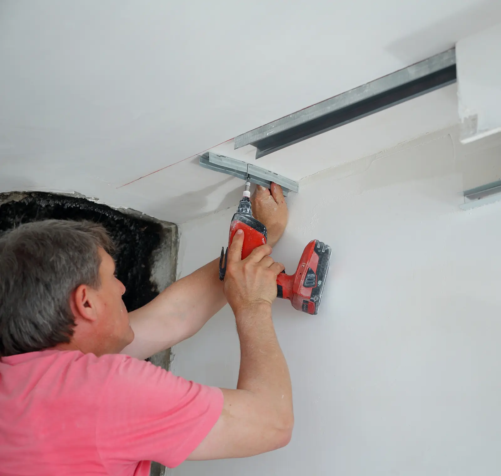 Person in a pink shirt uses a power drill to attach metal brackets to a white ceiling.