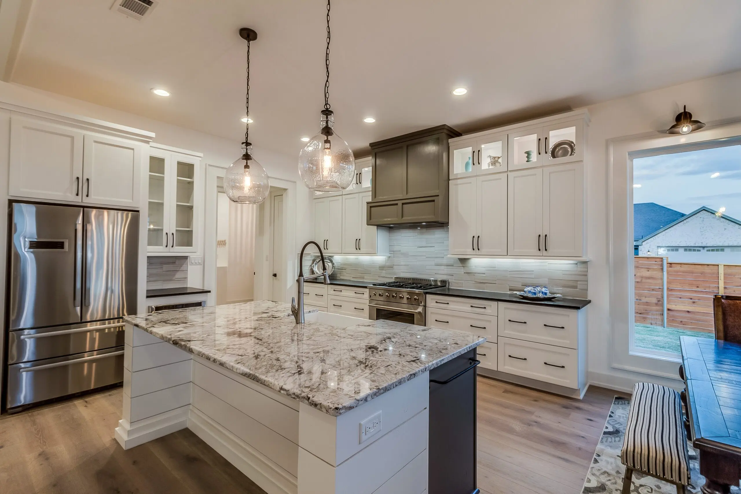 Modern kitchen with marble island, stainless steel appliances, white cabinets, and pendant lights. Large window on the right.